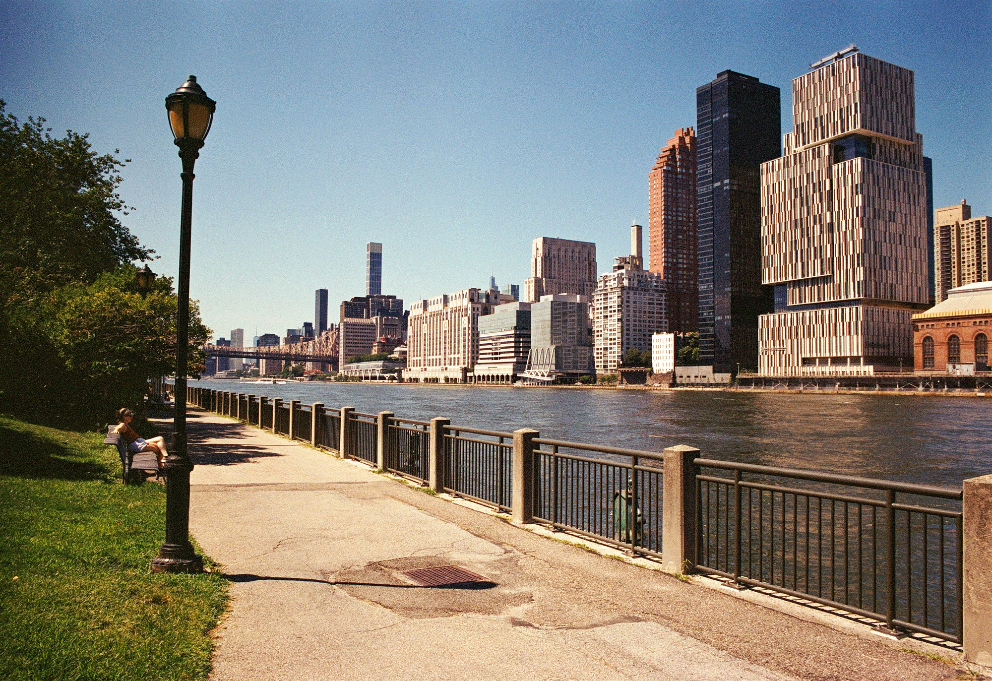 Roosevelt Island waterfront path with a seated visitor and the Manhattan skyline across the river