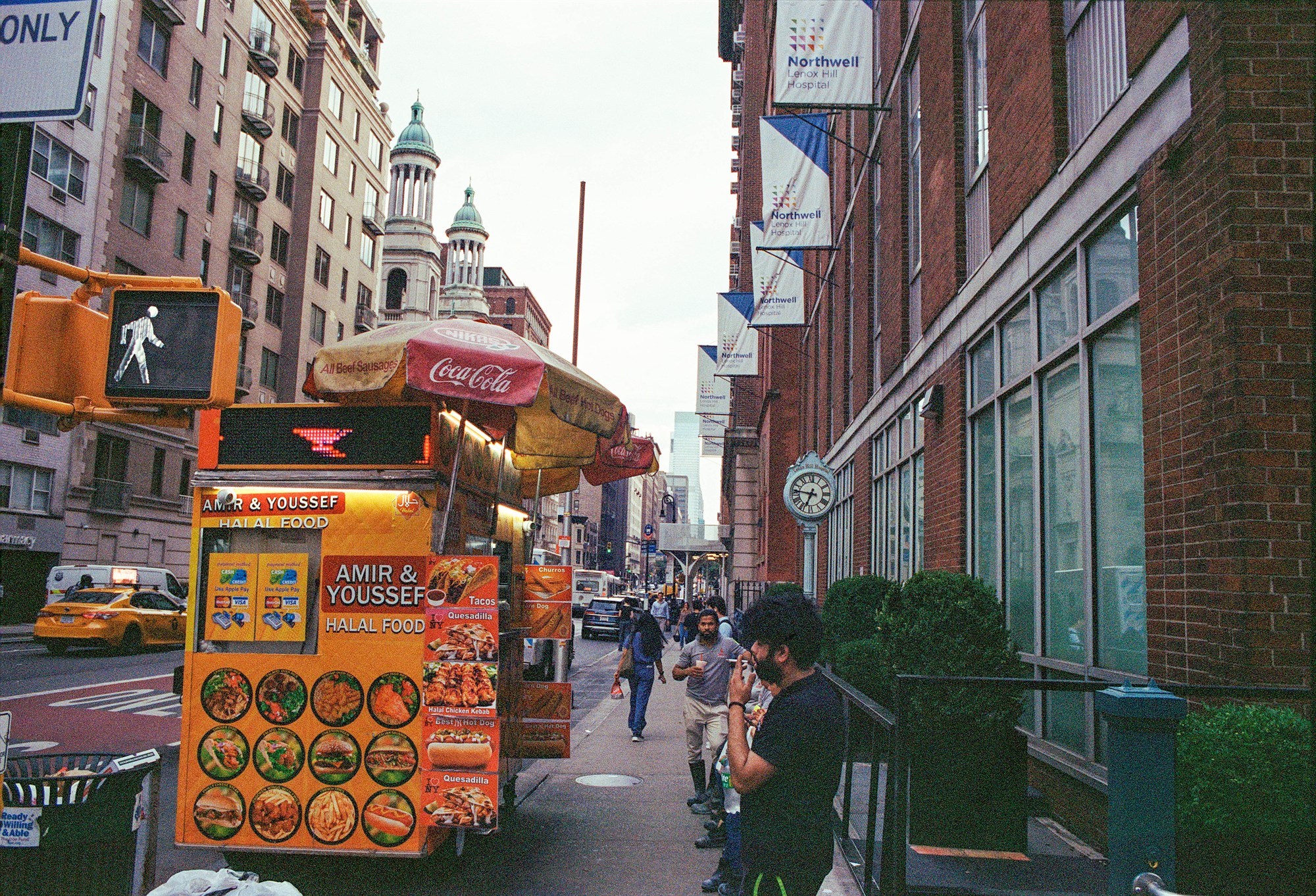 Busy halal food cart, pedestrians, and church towers along a Manhattan avenue